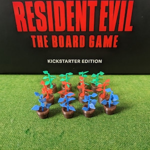 Könnte beinhalten: Ein Set mit 12 Miniatur-Topfpflanzen in verschiedenen Farben, darunter Grün, Rot und Blau. Die Pflanzen befinden sich in braunen Töpfen und sind in einer Reihe auf einer grünen Oberfläche angeordnet. Das Bild enthält den Text "Resident Evil The Board Game Kickstarter Edition".