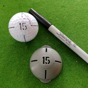 May include: A white golf ball with the number 15 written on it, a grey golf ball marker with the number 15 on it, and a black Sharpie permanent marker.