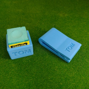 Puede incluir: Dos porta tizas azules con la palabra "TOM" impresa en ellos. Un porta tizas es una pequeña caja con una tiza azul dentro. El otro porta tizas es un estilo de clip.