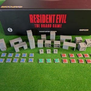 May include: A set of grey plastic miniature furniture pieces for a board game, including desks, cabinets, and shelves. The pieces are arranged on a green surface with a box in the background that reads "Resident Evil The Board Game Kickstarter Edition".