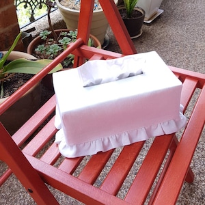 May include: A pink tissue box cover with white lace trim sits on a red wooden chair. The cover has a white tissue box insert.