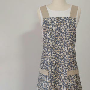 May include: A blue floral apron featuring small white and yellow flowers. It has tan straps and two tan pockets. The apron is displayed on a mannequin. The apron is designed for kitchen use.