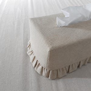 Puede incluir: Una funda rectangular para caja de pañuelos hecha de tela texturizada de color beige claro. La funda presenta un borde con volantes fruncidos en la parte inferior y una abertura rectangular en la parte superior, por donde sale un pañuelo blanco.