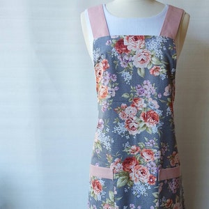 May include: A grey floral apron with pink straps and a large pocket. The apron has a floral pattern with pink, red and white flowers.
