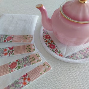 May include: Four white cotton tea towels with pink and floral patterned borders. A pink teapot with a gold trim sits on a white floral tea cosy.