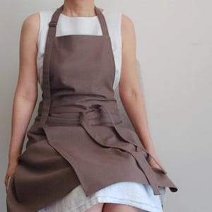May include: A brown apron with a bib and waist ties. The apron is made of a solid-colored fabric and has a simple, functional design. The apron is worn over a white top.