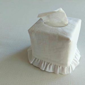 May include: A white linen tissue box cover with a ruffled edge. The square cover has a circular opening at the top for tissue dispensing. The design is simple and elegant, suitable for a bathroom or bedroom.