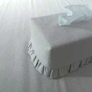 Puede incluir: Una funda rectangular para caja de pañuelos de tela beige claro con un ribete con volantes en el borde inferior. Los pañuelos blancos sobresalen de la abertura superior. La funda está sobre una superficie beige claro.