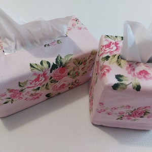 May include: Two pink tissue box covers with a floral pattern. The covers are made of fabric and have a square shape. The floral pattern features pink roses and green leaves.