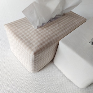 May include: Two rectangular tissue box covers. One cover features a beige and white gingham check pattern, while the other is a solid off-white colour. Both covers have a rectangular opening at the top with white tissues protruding from them.