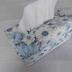 May include: A rectangular tissue box cover, decorated with a light blue floral design. The cover displays white flowers with blue centres and green leaves. White tissues are visible, protruding from the top opening. The box has a white trim.
