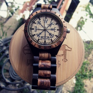 May include: A wooden wristwatch with a brown and black band. The watch face has a compass design with runes around the edge. The watch is on a wooden stand.