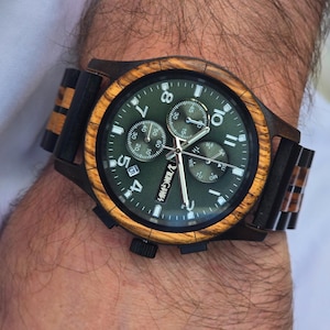 K&ouml;nnte beinhalten: Eine Holzarmbanduhr mit gr&uuml;nem Zifferblatt und brauner Holzl&uuml;nette und -armband. Das Zifferblatt hat wei&szlig;e Ziffern und Zeiger, mit dem Markennamen "KINGVINGS" sichtbar. Die Uhr befindet sich an einem Handgelenk.