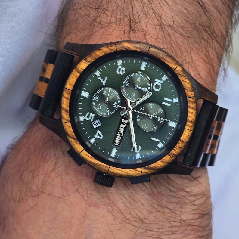 K&ouml;nnte beinhalten: Eine Holzarmbanduhr mit gr&uuml;nem Zifferblatt und brauner Holzl&uuml;nette und -armband. Das Zifferblatt hat wei&szlig;e Ziffern und Zeiger, mit dem Markennamen "KINGVINGS" sichtbar. Die Uhr befindet sich an einem Handgelenk.