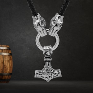 May include: A silver-colored pendant necklace featuring a detailed Mjolnir hammer design, suspended from a braided black leather cord. The necklace is connected by two wolf head clasps. The hammer is intricately engraved with Norse symbols.