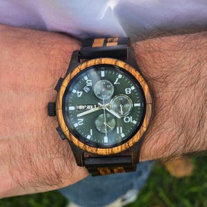 K&ouml;nnte beinhalten: Eine Holzarmbanduhr mit dunkelgr&uuml;nem Zifferblatt und Holzl&uuml;nette. Die Uhr hat ein schwarzes Armband mit Holzakzenten. Das Zifferblatt zeigt Zahlen und kleinere Zifferbl&auml;tter. Die Uhr ist an einem Handgelenk.