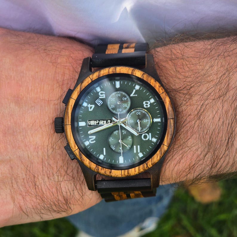 K&ouml;nnte beinhalten: Eine Holzarmbanduhr mit dunkelgr&uuml;nem Zifferblatt und Holzl&uuml;nette. Die Uhr hat ein schwarzes Armband mit Holzakzenten. Das Zifferblatt zeigt Zahlen und kleinere Zifferbl&auml;tter. Die Uhr ist an einem Handgelenk.