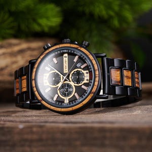 Puede incluir: Un reloj de pulsera negro con bisel y detalles de madera. La esfera del reloj es negra con subesferas y agujas doradas. La correa del reloj es negra con incrustaciones de madera. El reloj tiene el texto "VIKING".