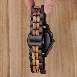 K&ouml;nnte beinhalten: Eine schwarze Holzuhr mit dunklem Zifferblatt und Holzarmband. Das Armband weist abwechselnd dunkle und helle Holzsegmente auf. Die Uhr wird vor einer hellbraunen Holzoberfl&auml;che gehalten.