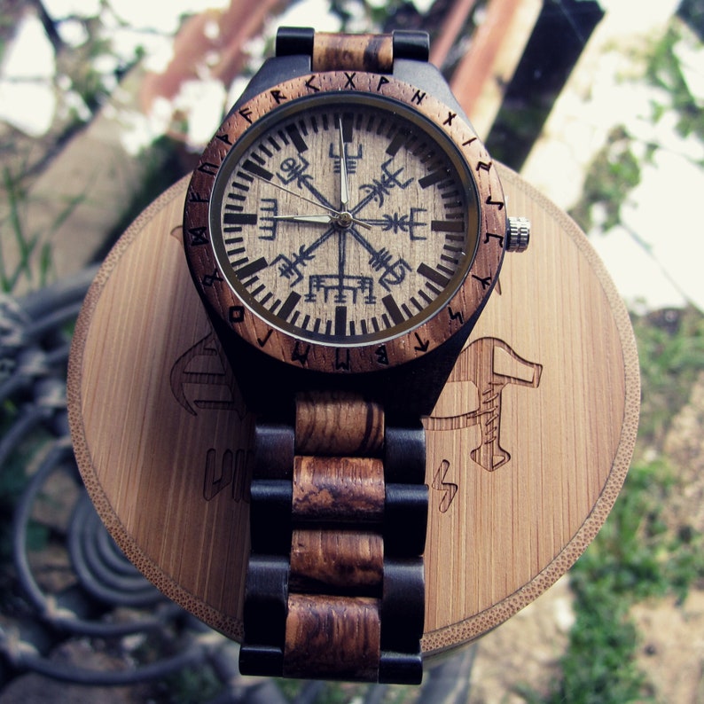 May include: A wooden wristwatch with a brown and black band. The watch face has a compass design with runes around the edge. The watch is on a wooden stand.