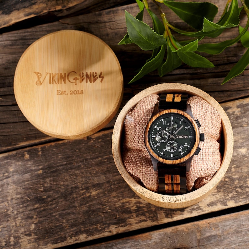 K&ouml;nnte beinhalten: Eine Holzuhr mit dunkelgr&uuml;nem Zifferblatt und Holzarmband wird in einer runden Bambusbox pr&auml;sentiert. Das Zifferblatt der Uhr hat silberfarbene Zeiger und Markierungen. Der Deckel der Box ist ge&ouml;ffnet und zeigt die Uhr und den Text "VIKING BNES Est. 2018".