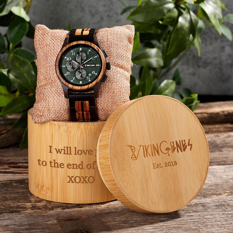 K&ouml;nnte beinhalten: Eine Holzuhr mit gr&uuml;nem Zifferblatt und braun-schwarzem Armband liegt auf einem kleinen Kissen. Die Uhr wird auf einem Bambusst&auml;nder mit der Aufschrift "I will love to the end of XOXO" pr&auml;sentiert. Daneben eine runde Bambusbox mit "VIKINGENES Est. 2018".