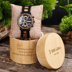 Puede incluir: Un reloj de madera con esfera negra y detalles dorados, exhibido sobre una almohada de arpillera dentro de una caja de bamb&uacute;. El reloj tiene una correa negra y con efecto madera. La caja est&aacute; grabada con texto. La tapa de la caja tambi&eacute;n es visible.