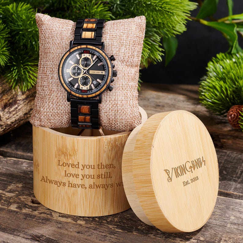 Puede incluir: Un reloj de madera con esfera negra y detalles dorados, exhibido sobre una almohada de arpillera dentro de una caja de bamb&uacute;. El reloj tiene una correa negra y con efecto madera. La caja est&aacute; grabada con texto. La tapa de la caja tambi&eacute;n es visible.