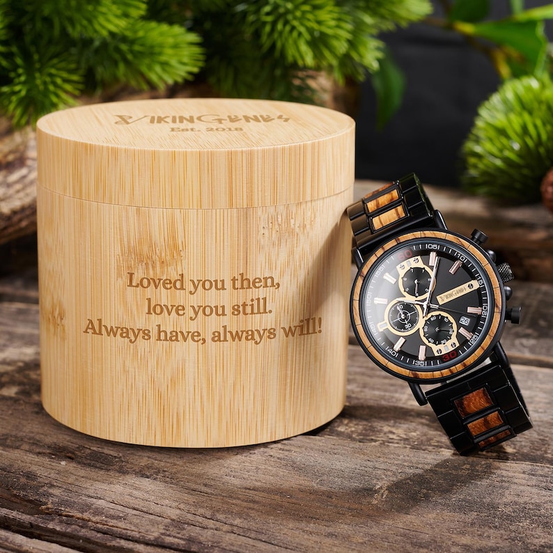 Puede incluir: Un reloj de madera con esfera negra y detalles en madera, junto a una caja cil&iacute;ndrica de madera. El reloj tiene tres subesferas y una ventana de fecha. La caja est&aacute; grabada con el texto "Loved you then, love you still. Always have, always will!"