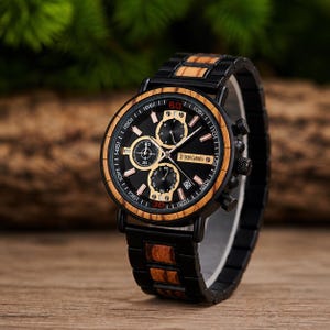 Puede incluir: Un reloj de pulsera negro con bisel y correa de madera. La esfera del reloj es negra con detalles dorados y tres subesferas. El reloj tiene el texto "KINGINFS" en la esfera. El reloj se muestra sobre una superficie de madera.