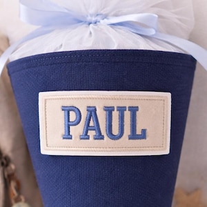 Könnte beinhalten: Eine dunkelblaue Schultüte aus Stoff mit einem beigen rechteckigen Aufnäher, auf dem der Name PAUL in blauem Garn gestickt ist. Die Öffnung ist mit weißem Tüll gerafft und mit einem hellblauen Band zu einer dekorativen Geschenktüte gebunden.