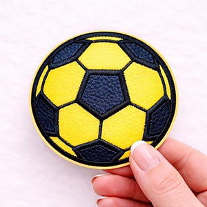 Könnte beinhalten: Ein runder Aufnäher, der einen gelben und marineblauen Fußball zeigt. Das Design zeigt das traditionelle Fünfeck- und Sechseckmuster mit schwarzen Nähten. Der Aufnäher wird von einer Hand gehalten, vor einem weißen Hintergrund.
