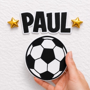 Könnte beinhalten: Ein runder, bestickter Aufnäher mit einem schwarz-weißen Fußballmotiv. Über dem Fußball befindet sich ein schwarzer, bestickter Namensaufnäher mit dem Namen PAUL in fetten Großbuchstaben, flankiert von zwei kleinen, dreidimensionalen goldenen Sternen.