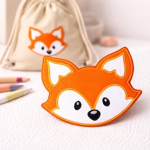 Könnte beinhalten: Ein orangefarbener Fuchs-Applikation mit weißen und schwarzen Details und einem gelben Umriss. Ein passendes Fuchsmotiv befindet sich auf einem beigen Kordelzugbeutel im Hintergrund. Farbstifte sind links zu sehen.