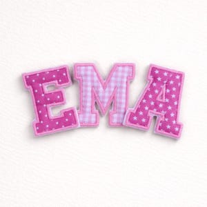Könnte beinhalten: Rosa Stoffbuchstaben mit der Aufschrift "EMA". Das "E" ist mit weißen Punkten bedeckt, das "M" hat ein rosa-weißes Vichy-Muster und das "A" hat weiße Sterne. Jeder Buchstabe hat einen rosa Rand.
