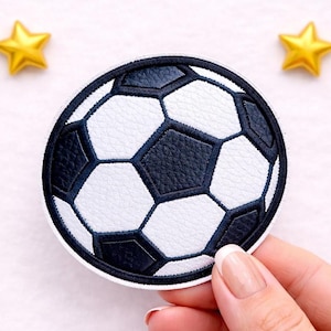 Könnte beinhalten: Ein runder Aufnäher mit einem Fußball-Design. Der Ball hat ein klassisches schwarz-weißes Muster mit strukturierter Oberfläche. Zwei goldene sternförmige Verzierungen befinden sich im Hintergrund.
