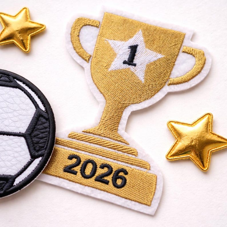 May include: An embroidered patch featuring a gold trophy with a white star containing the number 1, and the year 2026 in black text on the base. Beside it is a black and white soccer ball patch and two small, shiny gold star-shaped patches.