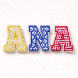 Könnte beinhalten: Drei gestickte Stoffbuchstaben mit der Aufschrift "ANA". Der Buchstabe "A" ist gelb mit einem Zickzackmuster, das "N" ist blau mit weißen Blumenmustern und das letzte "A" ist rot mit weißen Punkten.