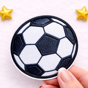 Könnte beinhalten: Ein runder Aufnäher, der einen Fußball zeigt, mit schwarzen und weißen Paneelen und detaillierten Nähten. Das Design zeigt das traditionelle Fußballmuster. Zwei goldene Stern-Dekorationen befinden sich im Hintergrund.