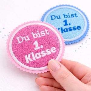 Könnte beinhalten: Zwei runde, glitzernde Abzeichen mit dem Text "Du bist 1. Klasse" in Weiß. Ein Abzeichen ist rosa mit einem gezackten Rand, das andere ist blau. Das rosa Abzeichen wird von einer Hand gehalten.