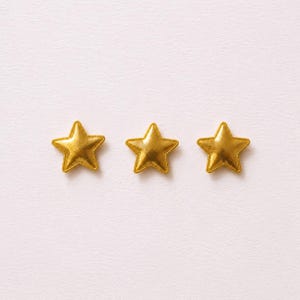 May include: Three small, five-pointed star-shaped embellishments with a metallic gold finish. Each star features a smooth, slightly raised surface with defined edges, arranged in a horizontal row against a plain, light-colored background.
