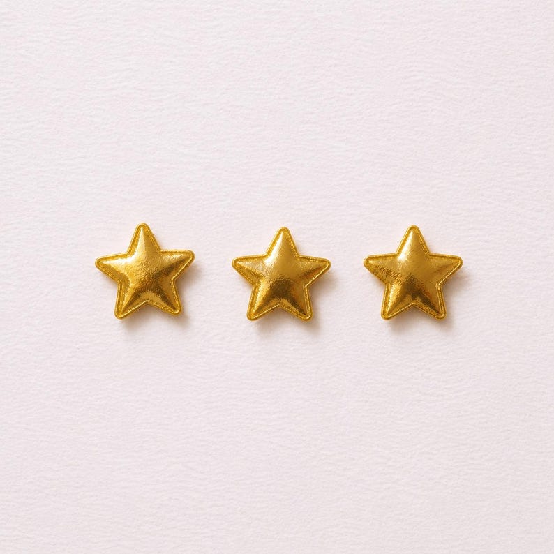 May include: Three small, five-pointed star-shaped embellishments with a metallic gold finish. Each star features a smooth, slightly raised surface with defined edges, arranged in a horizontal row against a plain, light-colored background.