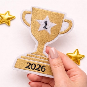 May include: A gold embroidered trophy patch with a white border, featuring a white star with the number 1 in the center and the year 2026 stitched in black on the base, accompanied by two small gold star accents on the sides of the trophy.