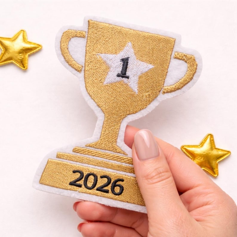 May include: A gold embroidered trophy patch with a white border, featuring a white star with the number 1 in the center and the year 2026 stitched in black on the base, accompanied by two small gold star accents on the sides of the trophy.