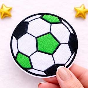 Könnte beinhalten: Ein runder, bestickter Aufnäher mit einem Fußballmotiv und strukturierter Oberfläche. Der Ball besteht aus weißen, schwarzen und hellgrünen fünfeckigen und sechseckigen Feldern, die mit dunklem Garn umrandet sind. Zwei kleine gelbe Sterne sind zu sehen.