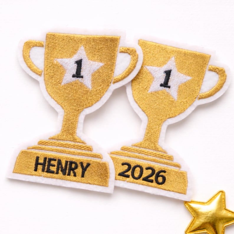 May include: Two gold trophy-shaped embroidered patches with white borders. Each trophy features a white star with a black number 1 in the center. The left patch has the name HENRY embroidered in black, and the right patch has the year 2026.