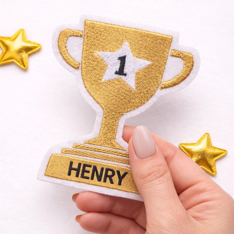 May include: A gold embroidered trophy patch with a white border, featuring a white star with the number 1 in the center and the name HENRY stitched in black on the base, accompanied by two small gold star accents on the sides.
