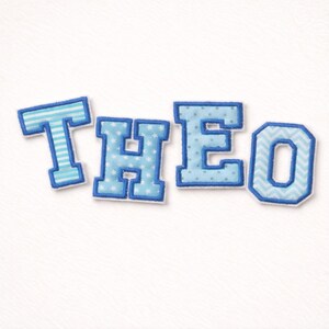Könnte beinhalten: Aufgenähte Stoffbuchstaben mit der Aufschrift "THEO" in Hellblau und Weiß. Jeder Buchstabe hat ein anderes Muster, darunter Streifen, Sterne, Punkte und ein Zickzack-Design. Die Buchstaben sind dunkelblau umrandet.
