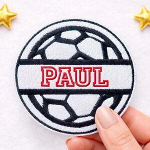 Könnte beinhalten: Runder Aufnäher mit einem Fußball-Design in Weiß und Schwarz, mit dem Namen "PAUL" in Rot gestickt. Der Name wird auf einem weißen Banner in der Mitte des Aufnähers angezeigt. Der Aufnäher wird von einer Hand gehalten.
