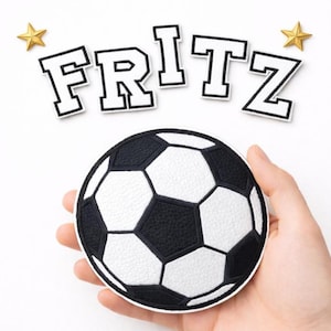 Könnte beinhalten: Ein runder Fußball-Aufnäher mit schwarz-weißem Strukturmuster, begleitet von einzelnen weißen Stoffbuchstaben mit schwarzem Rand, die das Wort FRITZ bilden, sowie zwei kleinen goldfarbenen sternförmigen Verzierungen.